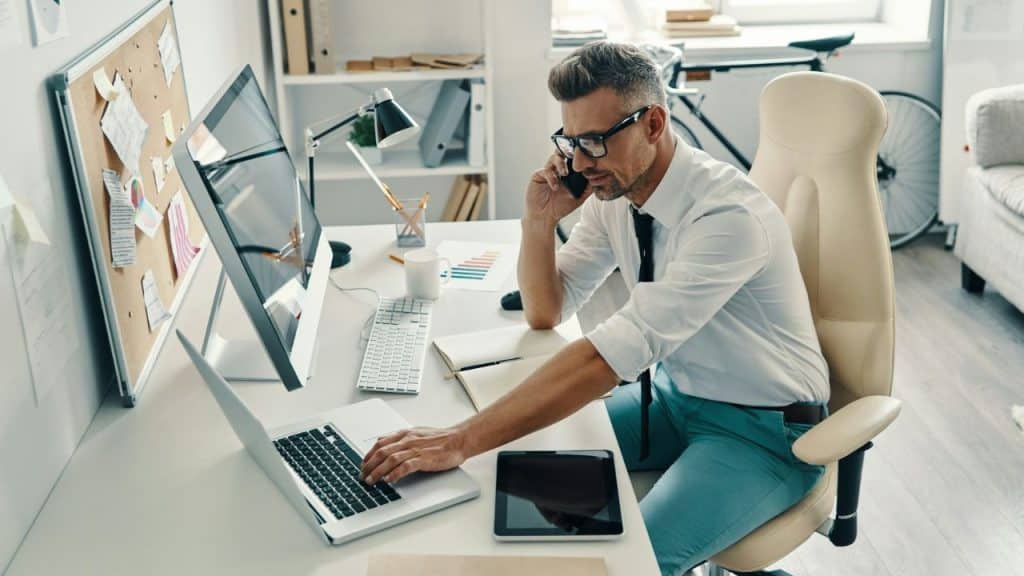 Middle-aged man on a phone call while typing on a laptop in a bright office.
