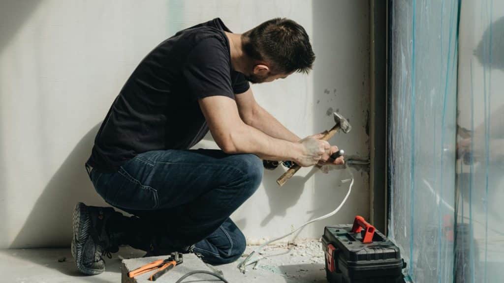 Man squatting, hammering near a wire coming from the wall by a window.