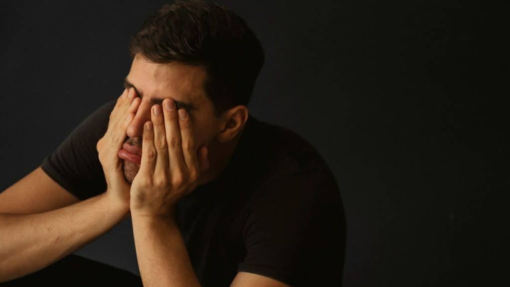 Man wearing a dark shirt covering his eyes with both hands against a dark background.