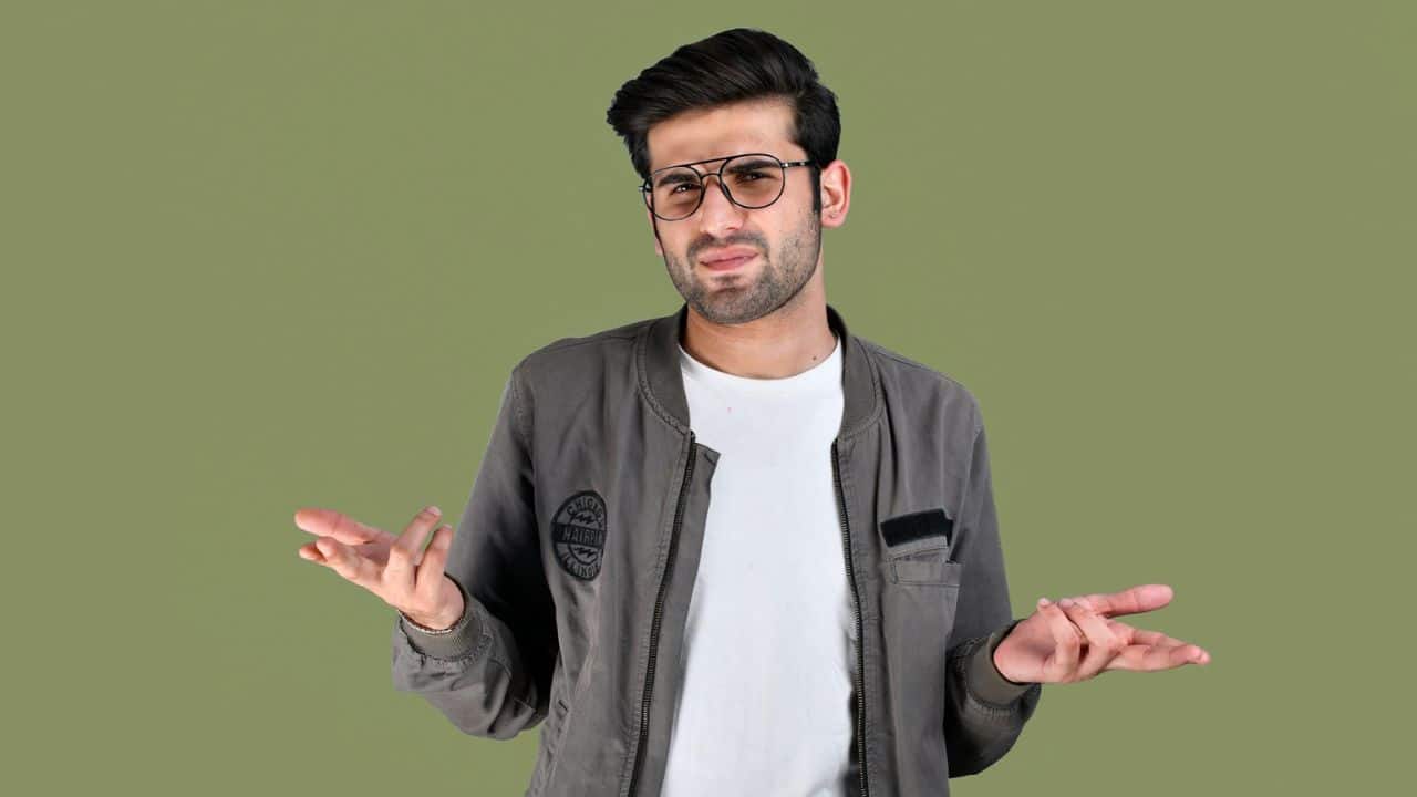 A man in a bomber jacket and glasses shrugging with open hands against a green background.