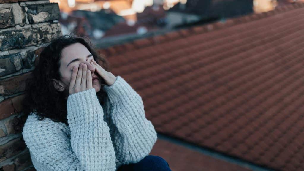 A person wearing a knitted sweater sits by a brick wall, covering their face with their hands.