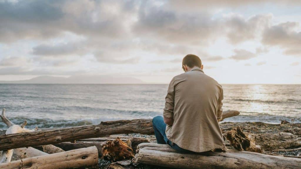 A man sitting on driftwood by the beach, looking at the ocean during sunset.