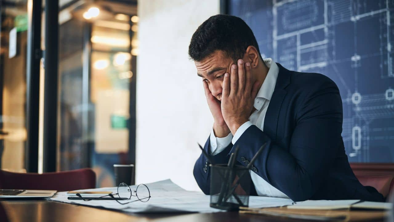 Man in a suit sits at a desk with his head in his hands, looking distressed.
