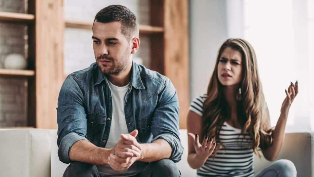 Man looking away with hands clasped as a woman gestures angrily in the background.
