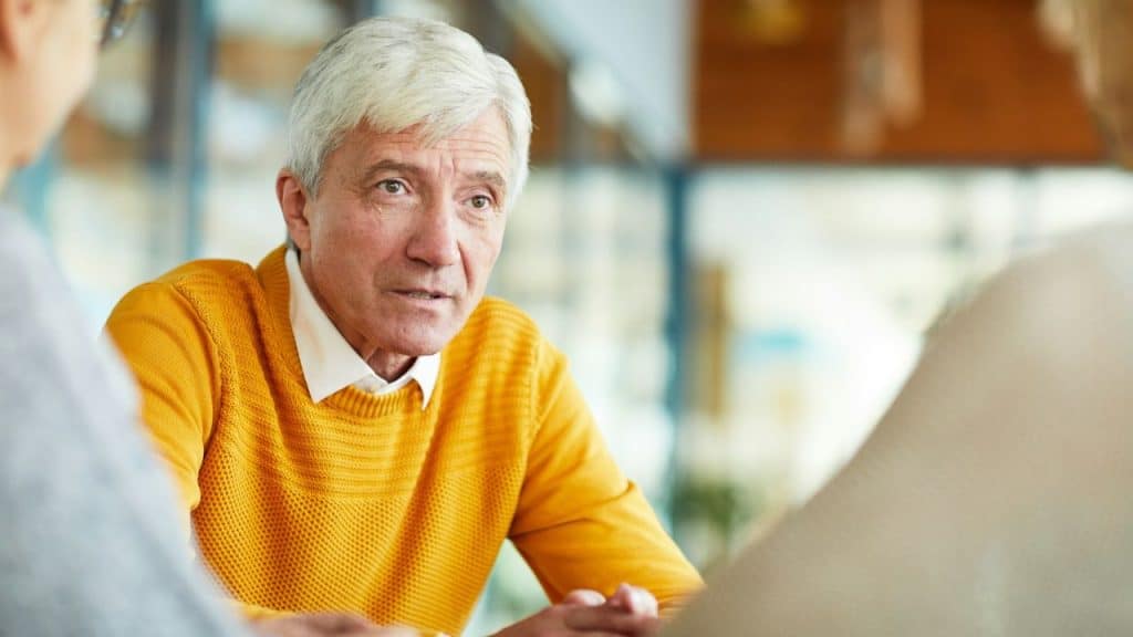 Older man in a bright yellow sweater talking intently to people across from him.