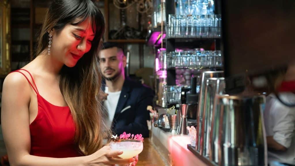 A woman in a red dress holding a cocktail at a bar with a man in the background.