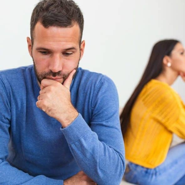 A thoughtful man in blue sits in the foreground with a woman blurred behind him.