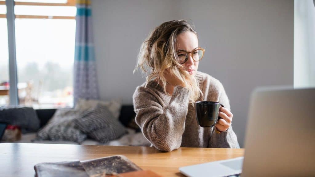 A woman wearing glasses sits at a table with a laptop, holding a mug and appearing thoughtful.
