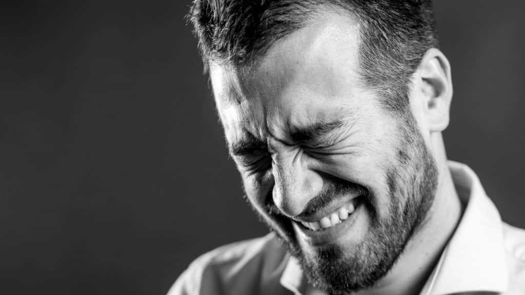 A black and white close-up of a bearded man with his eyes tightly closed, showing an expression of intense emotion.
