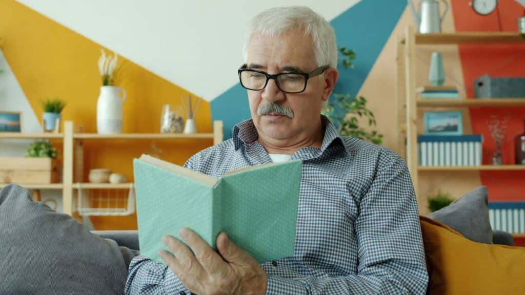 Older man with glasses and a mustache reading a book in a colorful living room.