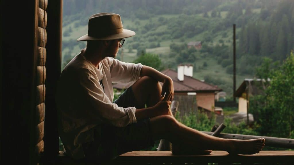 Man in a hat and glasses sitting thoughtfully, looking out at a hilly, green landscape.