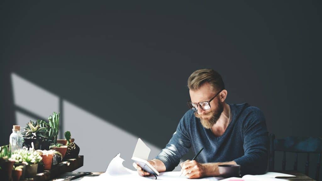A focused man sitting at a table is writing something on a paper.
