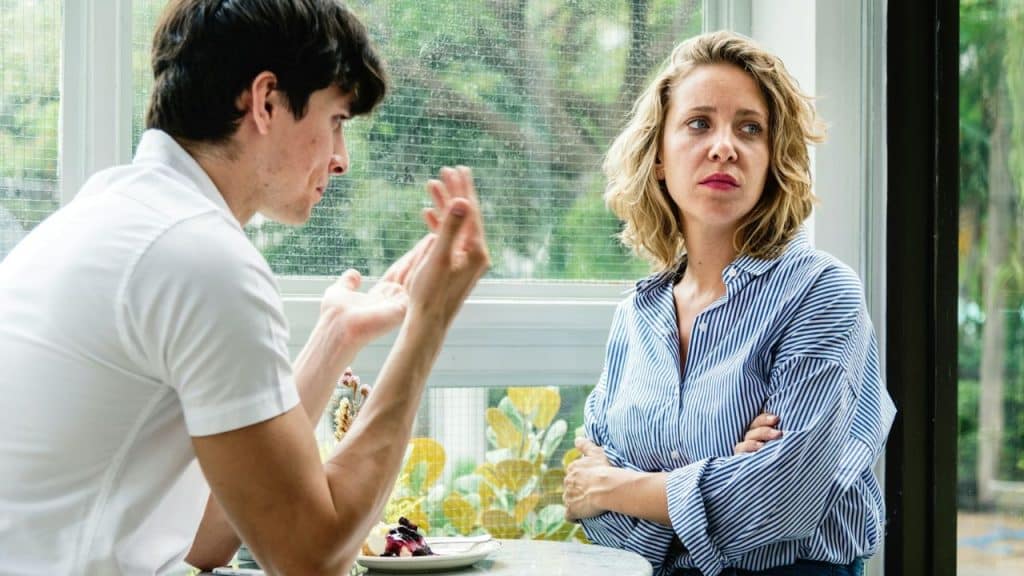 A man talks emphatically across a table to a woman who is looking away with crossed arms.