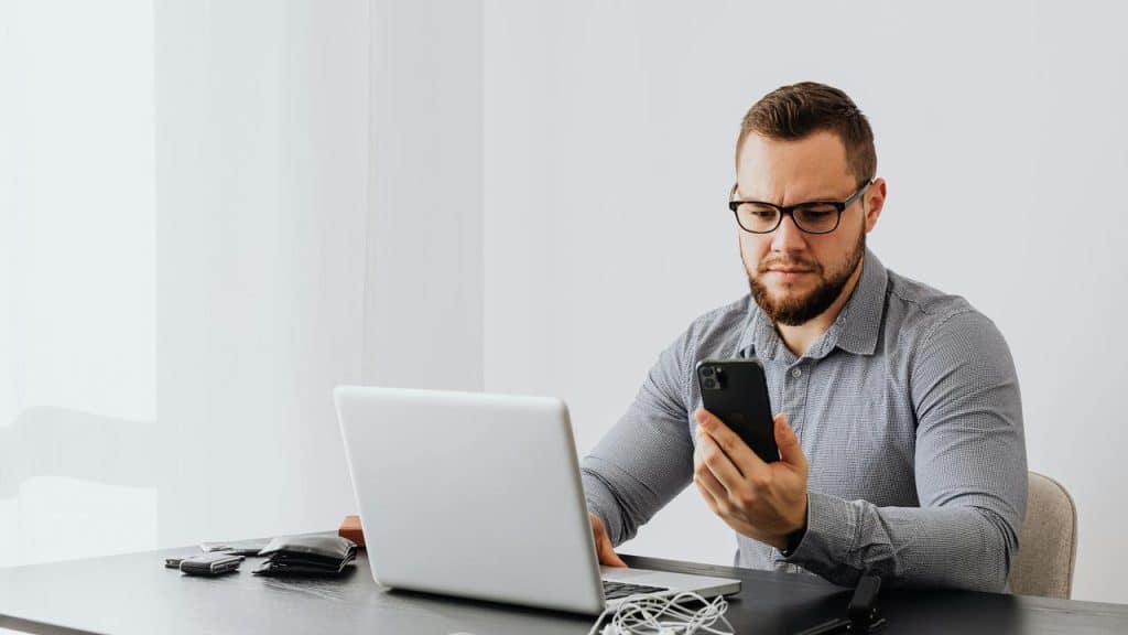 A man sitting at a desk looking at his phone with a laptop in front of him.