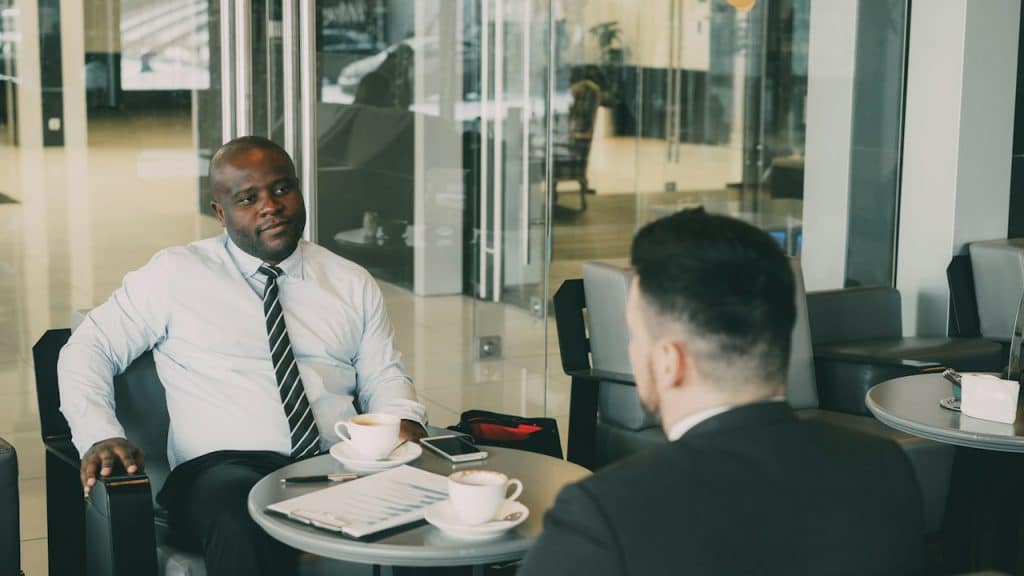 A bald man looking at another man while having coffee as they sit in an office.