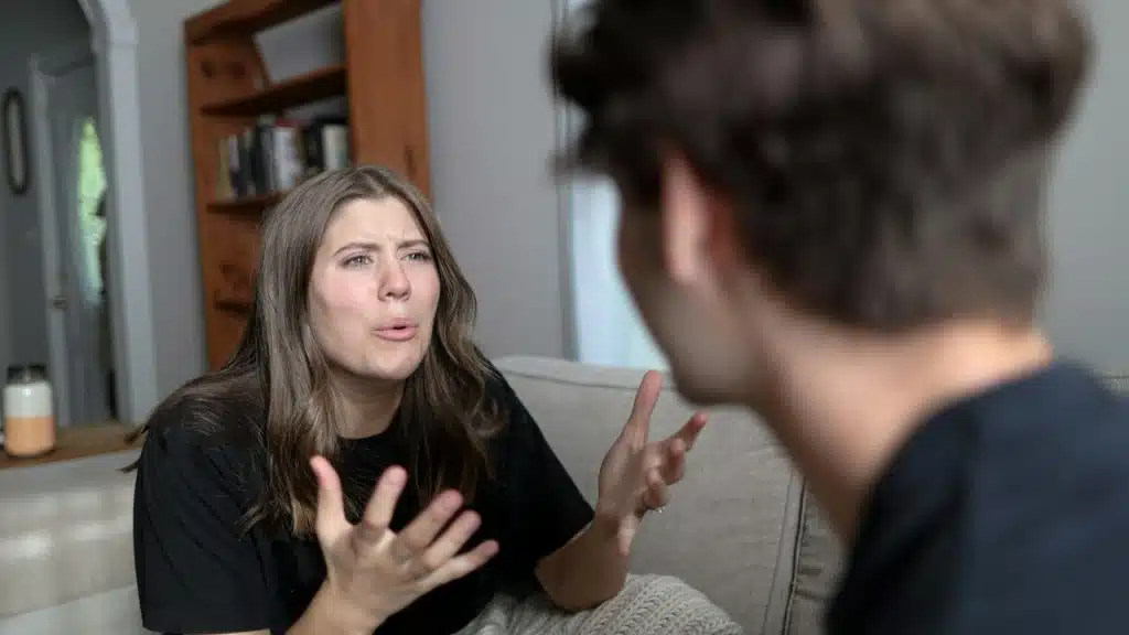 A woman looking distressed and gesturing at a man while they sit on a couch in a living room.