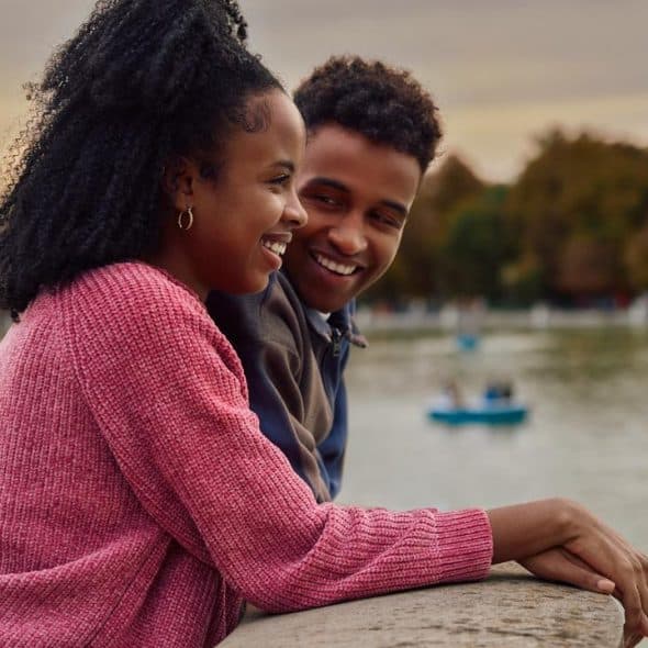A smiling couple leans on a lakeside railing together at sunset.