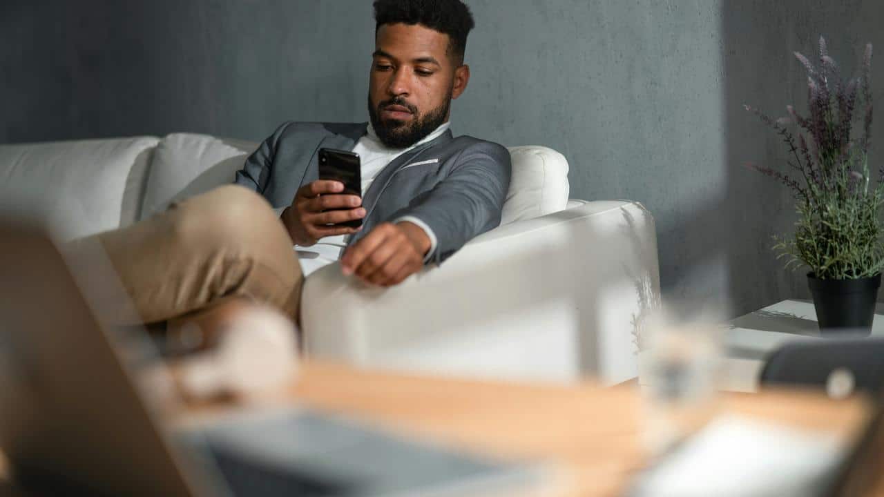 A man in a blazer sitting on a couch while using his phone.