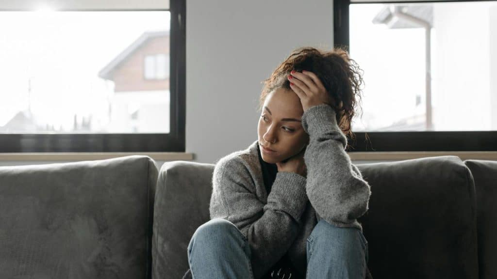 A woman sits on a couch looking upset with her hand on her head.