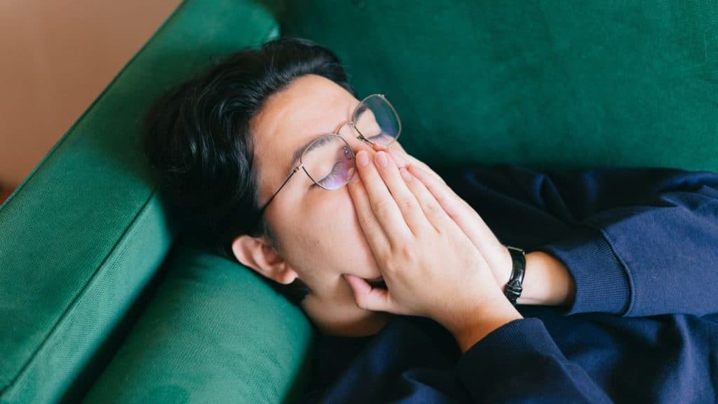A person with glasses lies on a green couch, covering their mouth and nose with their hands.