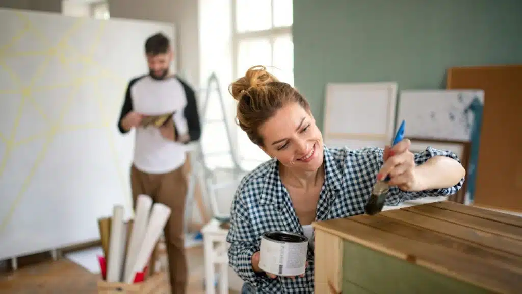 A smiling woman paints a wooden crate while a man works on a wall in the background.