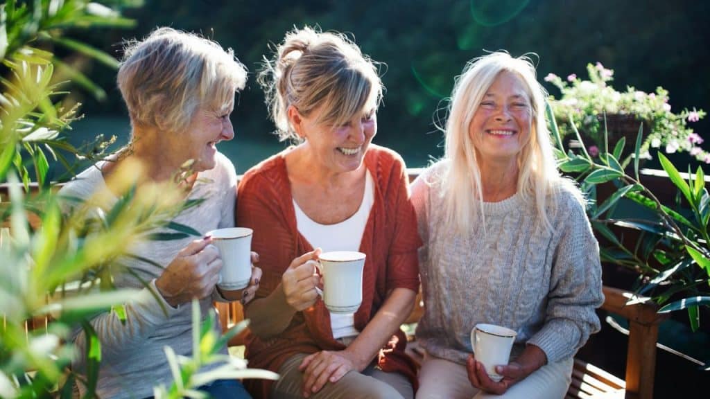 Three smiling older women sit outdoors, drinking from mugs and laughing together.