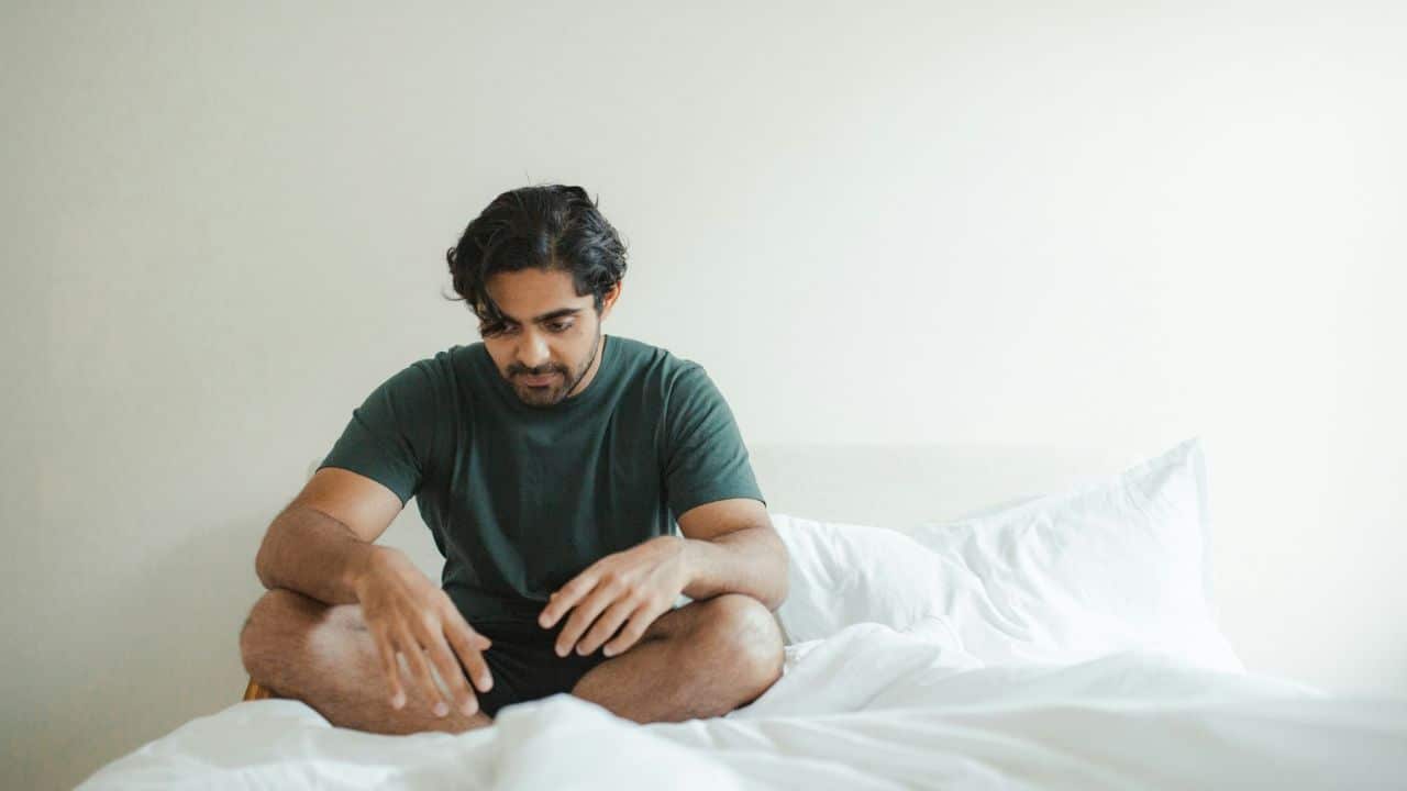 A man with dark hair sits cross-legged on a white bed, looking down with a somber expression.
