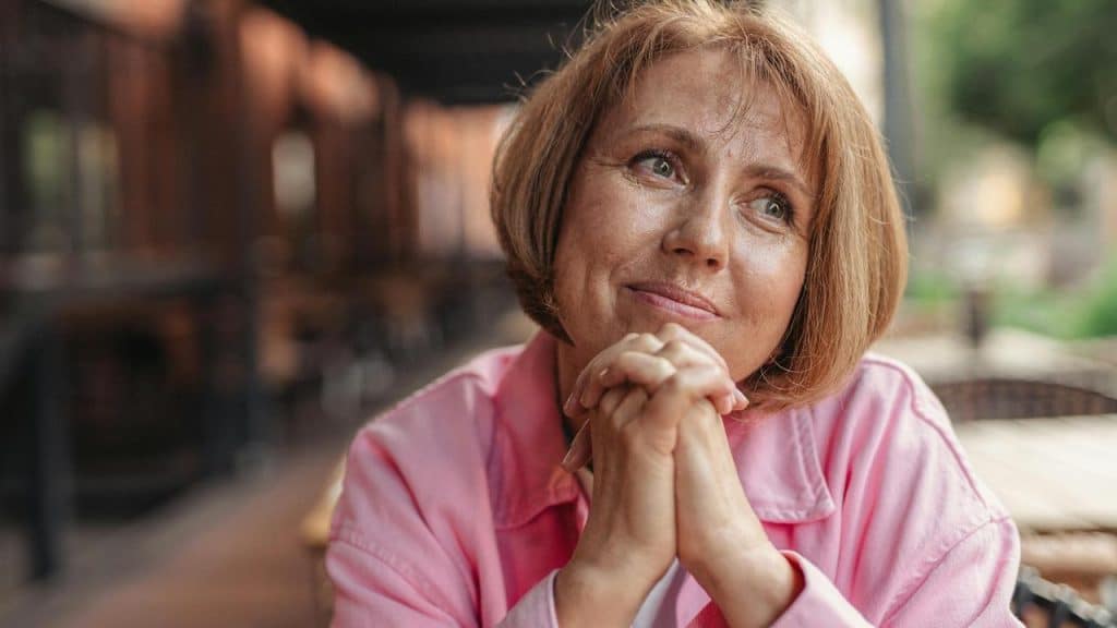 A woman in a pink shirt sits outdoors with her hands clasped, smiling softly.
