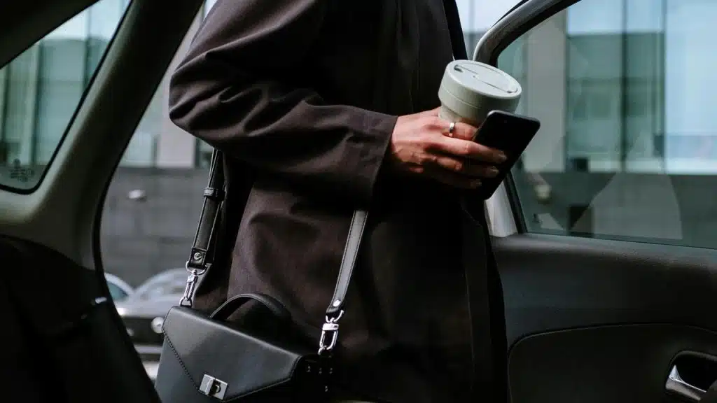 A person holding a phone and a reusable cup while getting into a car.