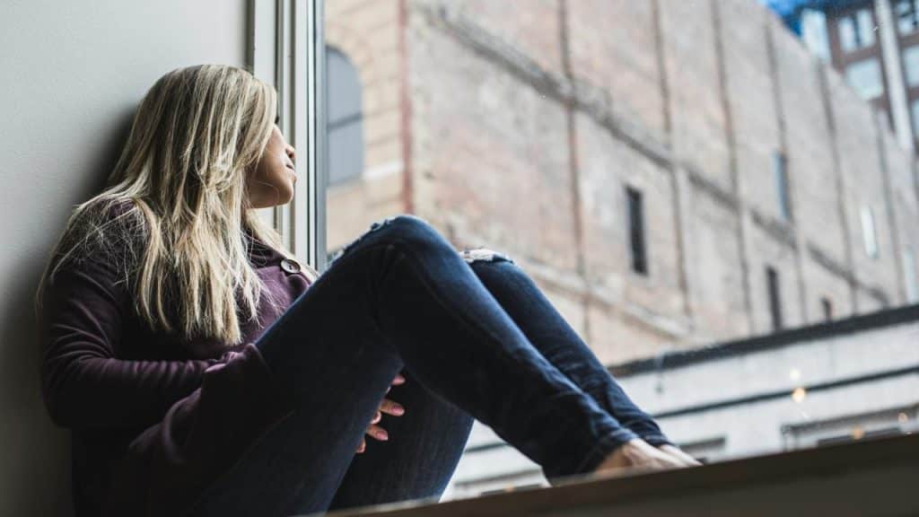 A woman sits by a window, gazing outside at a brick building.