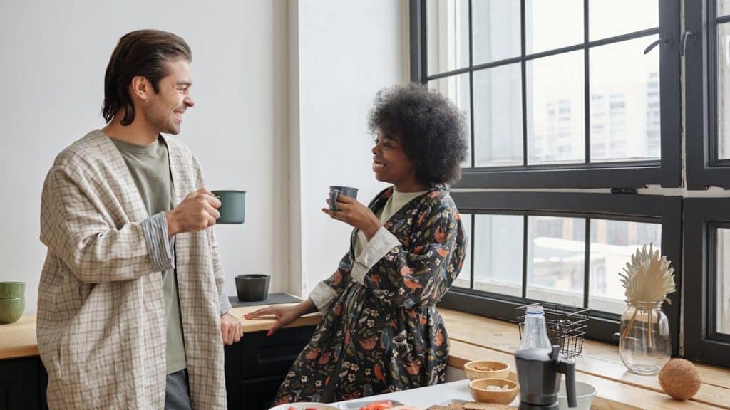 Smiling couple in robes enjoying coffee and talking in a bright kitchen with breakfast.