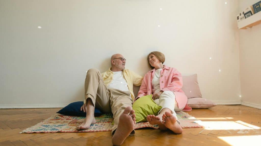 A senior couple sitting on the floor against a wall, smiling at each other.