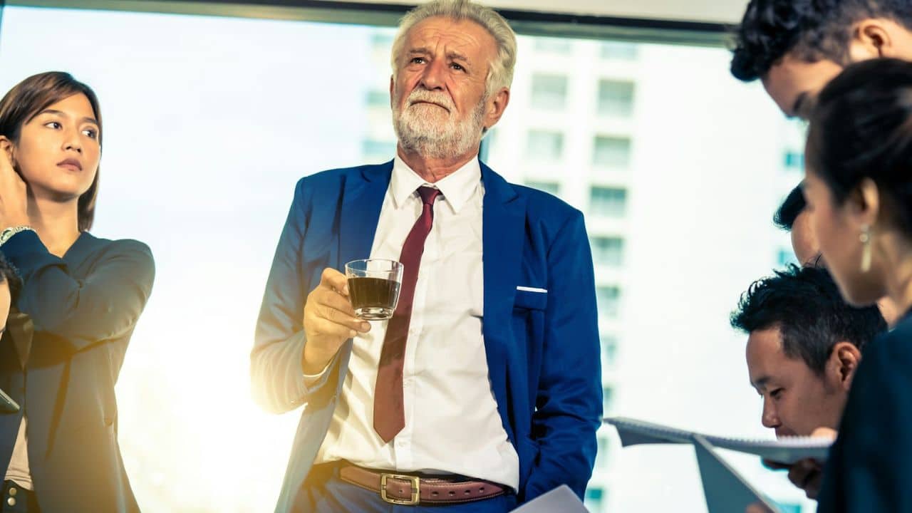 A gray-bearded man in a blue suit holds a drink, surrounded by colleagues in an office.