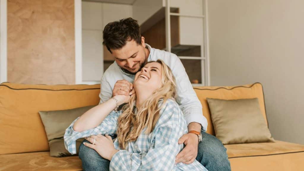 A couple laughing together while sitting on a couch in a cozy living room.