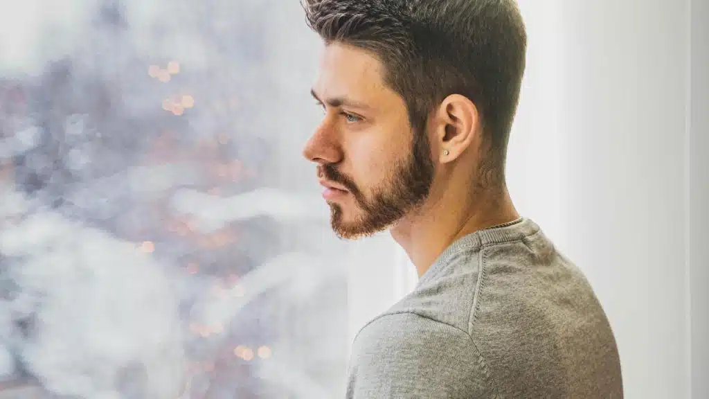 A man with a trimmed beard looking pensively out a bright window.