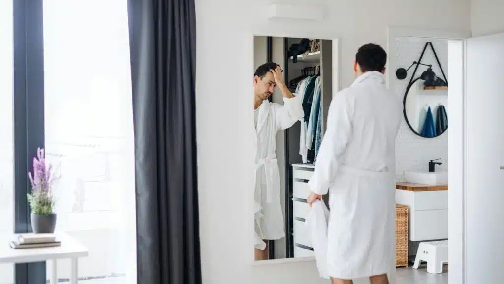 A man in a white bathrobe is standing in front of a mirror in a bright room, adjusting his hair.
