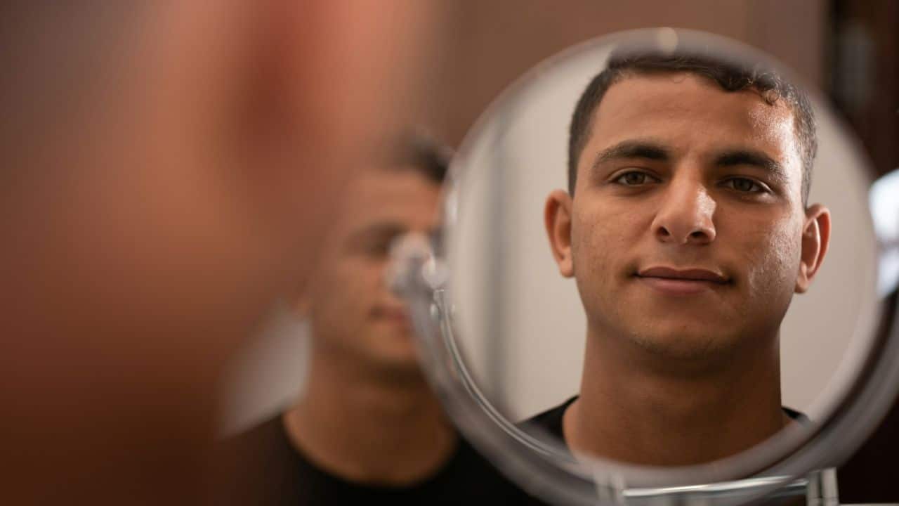A young man's face is reflected in a handheld magnifying mirror.