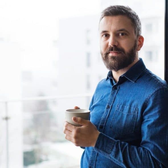 A bearded man in a blue shirt holds a mug on a balcony overlooking white buildings.