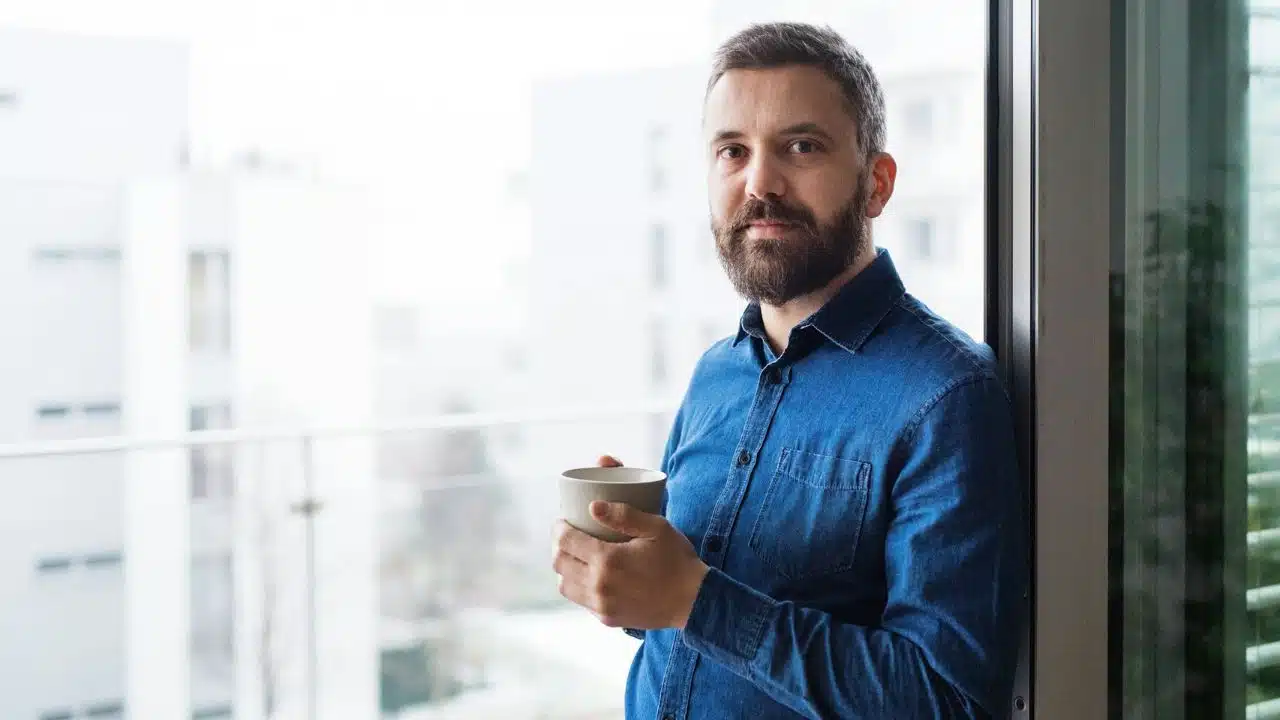 A bearded man in a blue shirt holds a mug on a balcony overlooking white buildings.