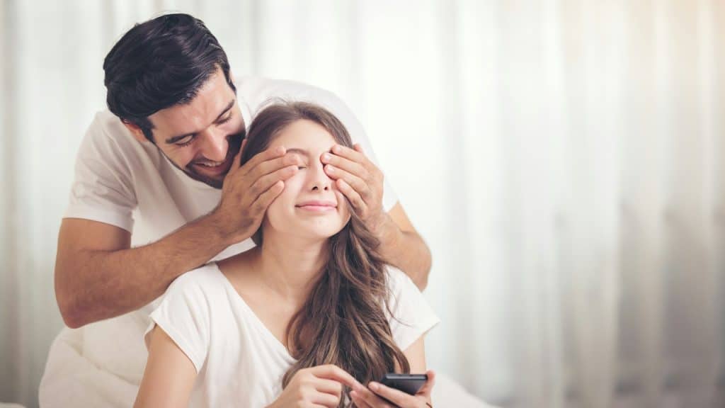 A smiling man covering his wife’s eye.
