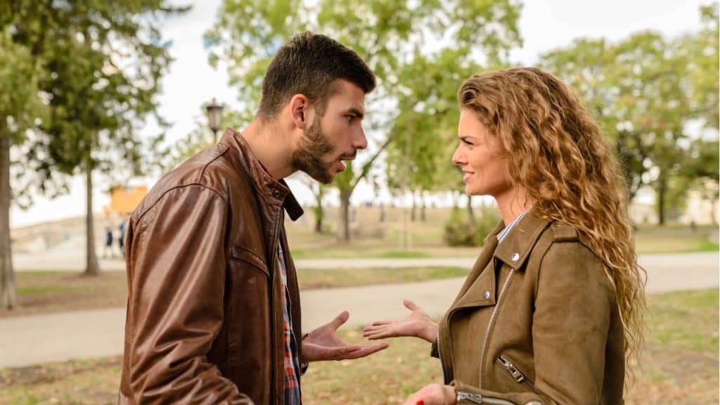 A man and a woman in leather jackets are standing outdoors, facing each other and arguing.