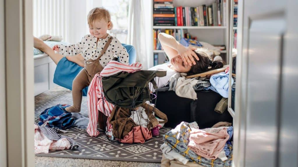 A tired woman is overwhelmed by a pile of laundry while a baby plays nearby.