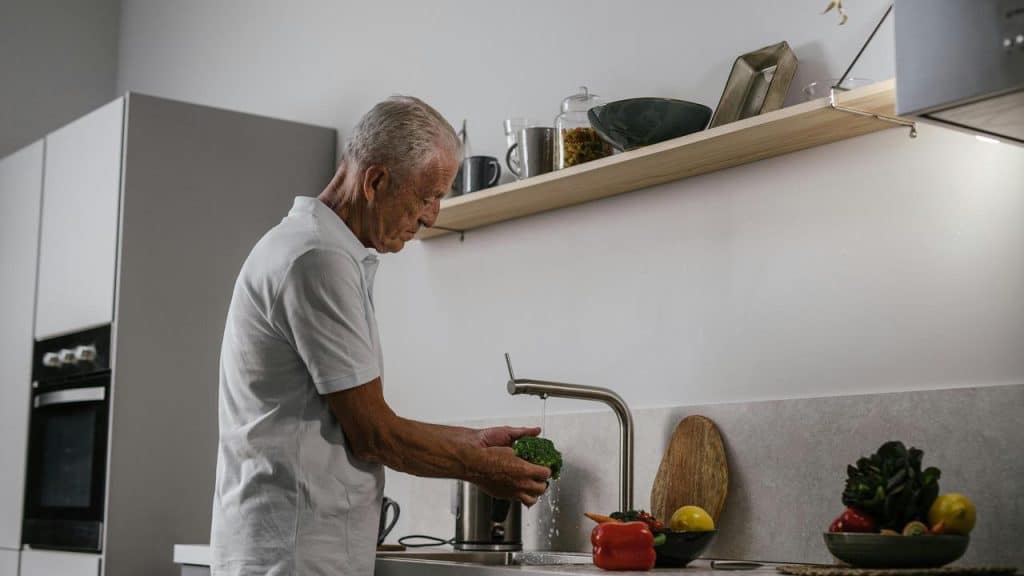 An elderly man washing vegetables at a kitchen sink.