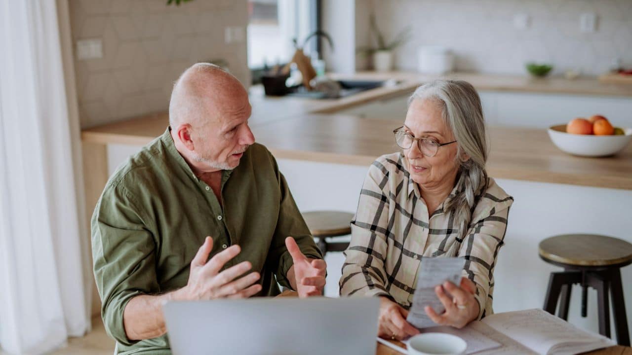 An elderly couple sits at a table with a laptop, discussing papers in a kitchen.