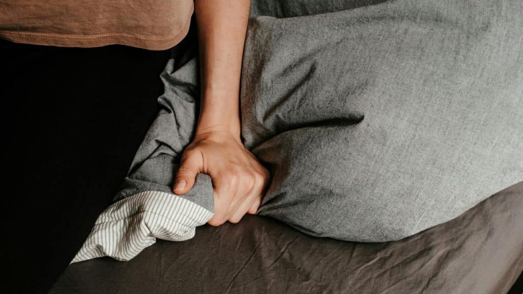 A person gripping a gray blanket tightly on a bed.
