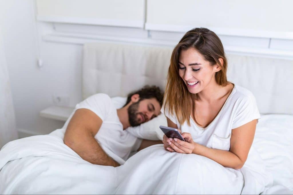 A woman busy with her [hone and a man sleeping beside her