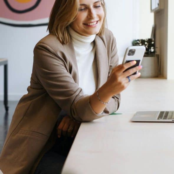 A woman sitting at a long desk, smiling at her phone beside an open laptop.