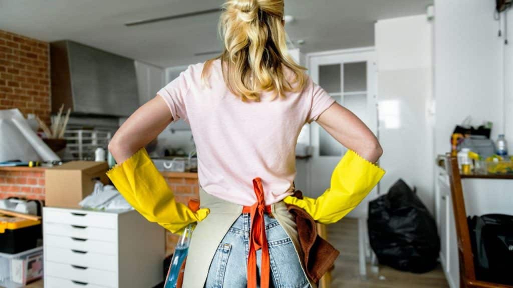 A person with blonde hair stands with hands on hips, wearing yellow gloves and an apron.