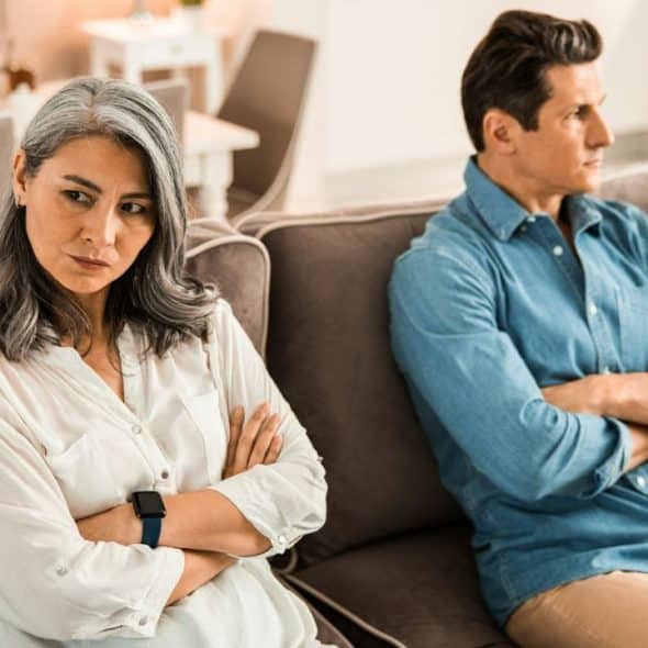 Woman with gray hair and man in denim shirt sit on a sofa with crossed arms, looking away.