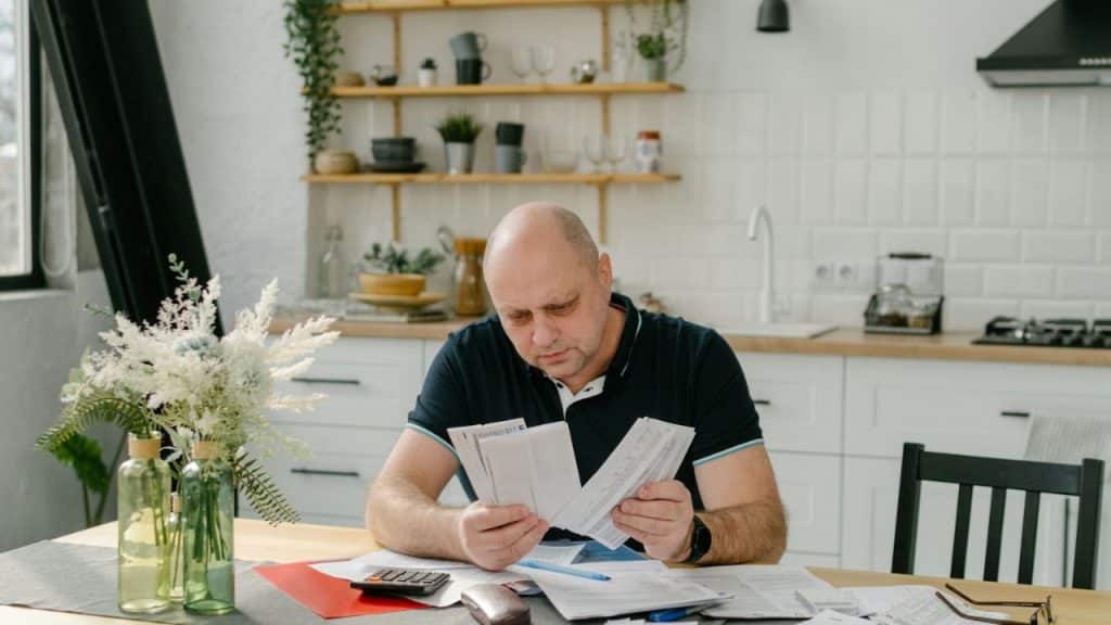 Bald man in a dark shirt reviews documents at a kitchen table.